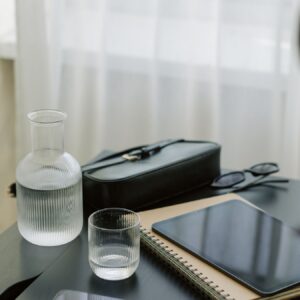 Minimalistyczne biurko z notatnikiem, tabletem, karafką z wodą i szklanką, eleganckim etui oraz okularami, na tle jasnych firan.